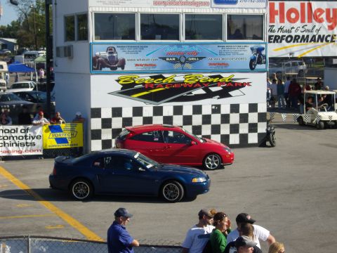 2007 Ford Mustang - Two cars are parked at a raceway with a tower and signage in the background.