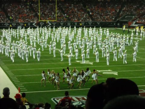 1979 Ford F-150 - A large marching band and cheerleaders are on a football field, with spectators in the background.