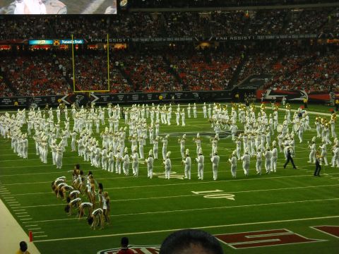 1979 Ford F-150 - A marching band performs on a football field during a game, with fans filling the stadium seats.