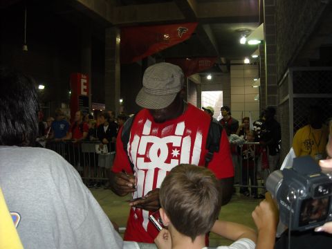 1979 Ford F-150 - A man wearing a red shirt with a large white DC logo signs autographs for fans while a crowd watches.