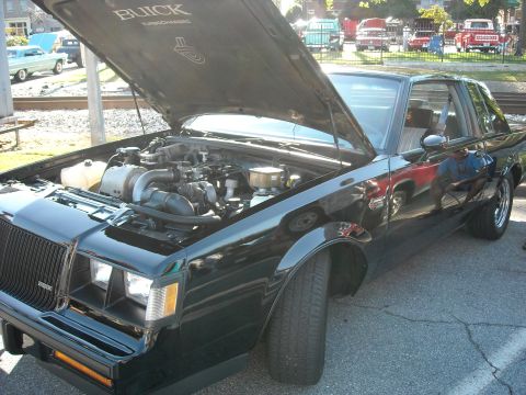 1979 Ford F-150 - A black Buick Grand National is parked with its hood up, revealing the engine bay. Several other vehicles are visible in the background.