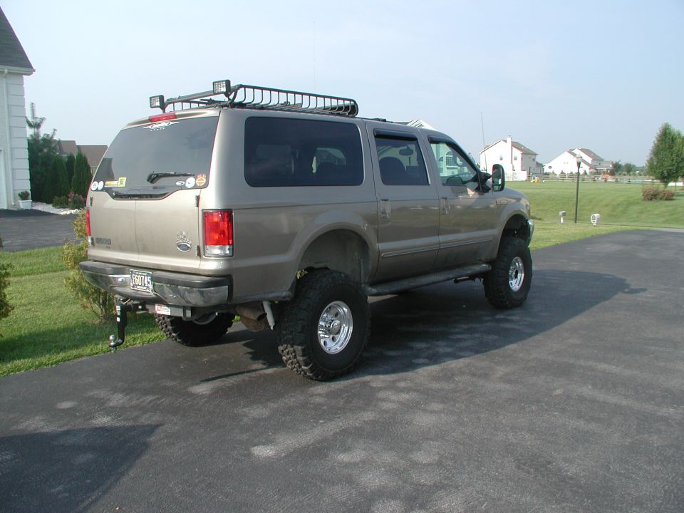 Gold lifted Ford Excursion with roof rack, rear three-quarter view in a suburban driveway