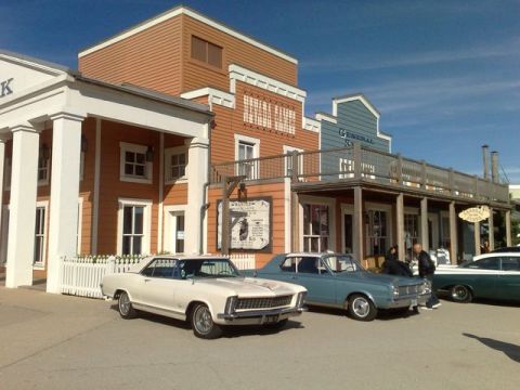 1991 Ford LTD Crown Victoria - A white 1965 Buick Riviera coupe is parked in front of a western-themed building with other vintage cars.