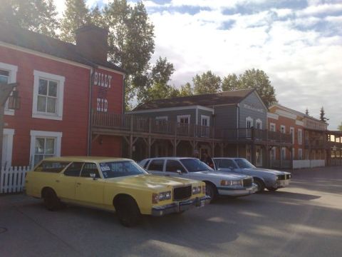 1991 Ford LTD Crown Victoria - A yellow 1977 Ford Country Squire station wagon is parked alongside other vehicles in front of a row of old west-style buildings.