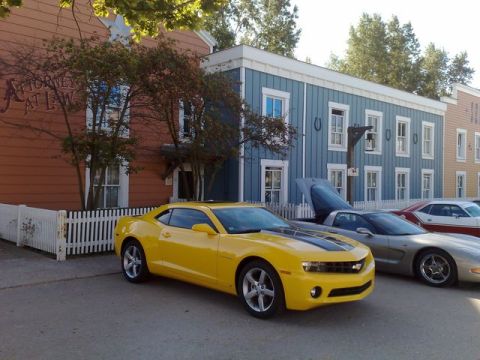 1991 Ford LTD Crown Victoria - A yellow Chevrolet Camaro and a silver Chevrolet Corvette are parked in front of buildings with a vintage facade.