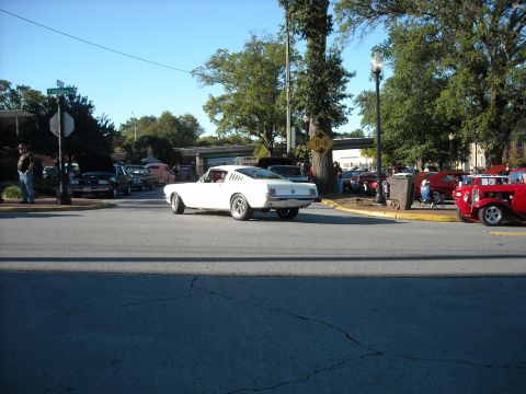 1979 Ford F-150 - A white classic coupe is seen from the rear, driving past a gathering of other vehicles on a sunny day.