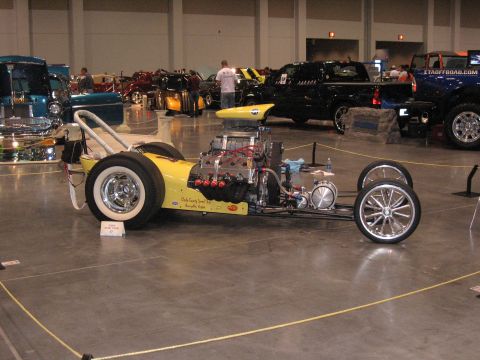 1968 Shelby GT500 - A yellow dragster with a prominent engine is displayed indoors, with other vehicles in the background.