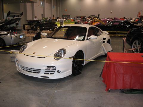 1968 Shelby GT500 - A white Porsche 911 Turbo is displayed indoors at a car show, with other vehicles visible in the background.