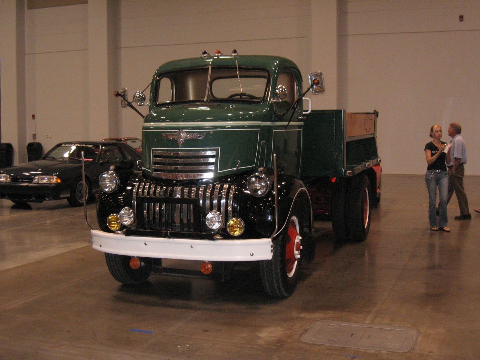 Green and black flatbed truck with chrome grille and white bumper. Black car in background