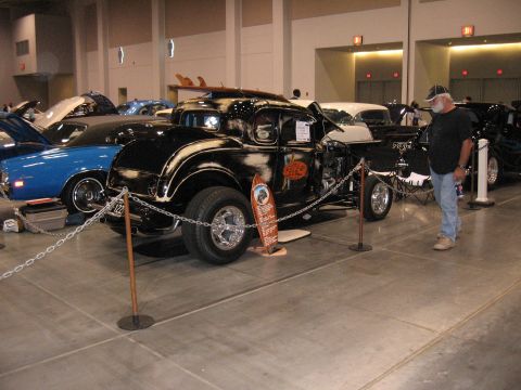 1968 Shelby GT500 - A man observes a custom hot rod coupe with surfboards on its roof at an indoor car show.