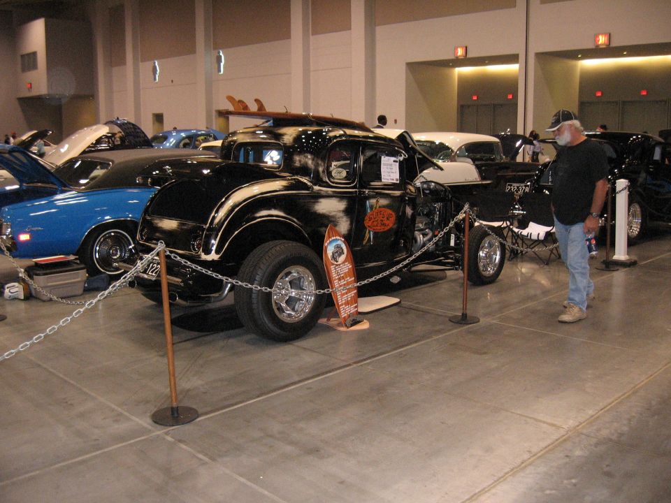 Black hot rod coupe with surfboards on roof, chrome wheels, and custom paint, displayed indoors