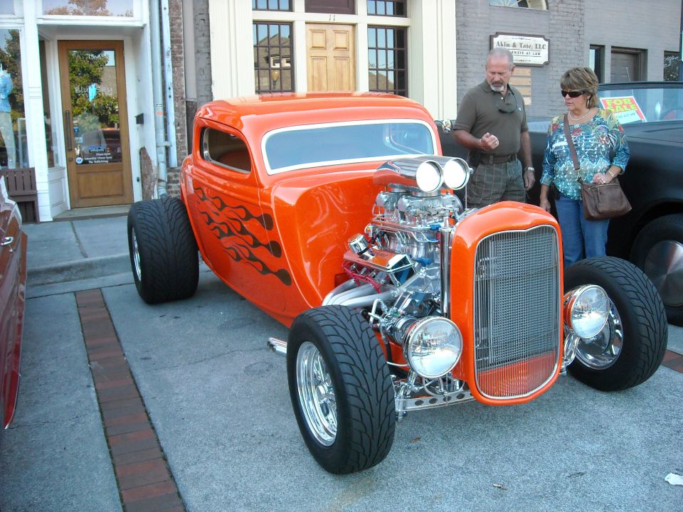 Orange hot rod with flame decals and a large supercharged engine. Two people stand nearby