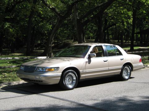 2000 Mercury Grand Marquis - A tan sedan is parked on the side of a road, with trees and foliage in the background.