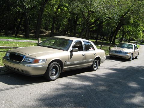 2000 Mercury Grand Marquis - Two gold Mercury Grand Marquis sedans are parked on a tree-lined road.