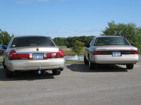 2000 Mercury Grand Marquis - Two Mercury Grand Marquis sedans are parked near a body of water with trees in the background under a clear blue sky.
