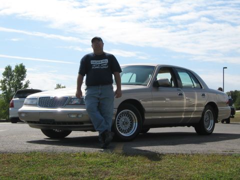 2000 Mercury Grand Marquis - A man stands next to a tan 2000 Mercury Grand Marquis at an outdoor event.