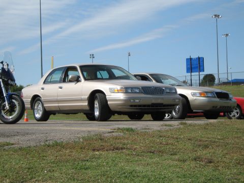 2000 Mercury Grand Marquis - Two Mercury Grand Marquis sedans are parked side-by-side on a sunny day, with a motorcycle to the left.
