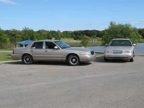 2000 Mercury Grand Marquis - Two Mercury Grand Marquis sedans are parked outdoors near a lake on a sunny day.