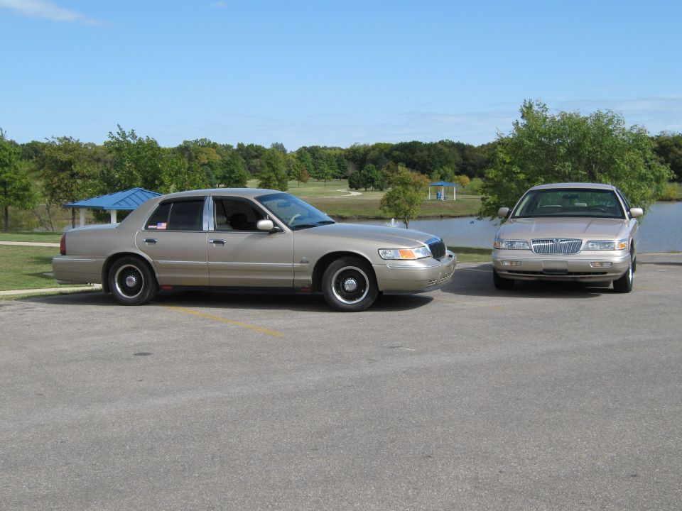 Two tan 2000 Mercury Grand Marquis sedans parked side-by-side on asphalt with trees and a lake in the background