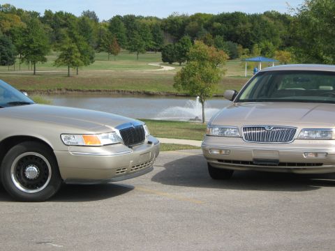 2000 Mercury Grand Marquis - Two similar Mercury Grand Marquis sedans are parked side-by-side in a lot with a pond and trees in the background.