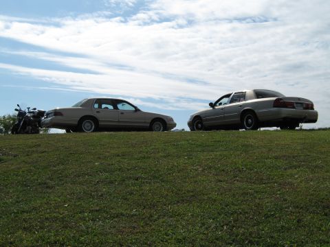 2000 Mercury Grand Marquis - Two sedans are parked on a grassy incline with a cloudy sky above.