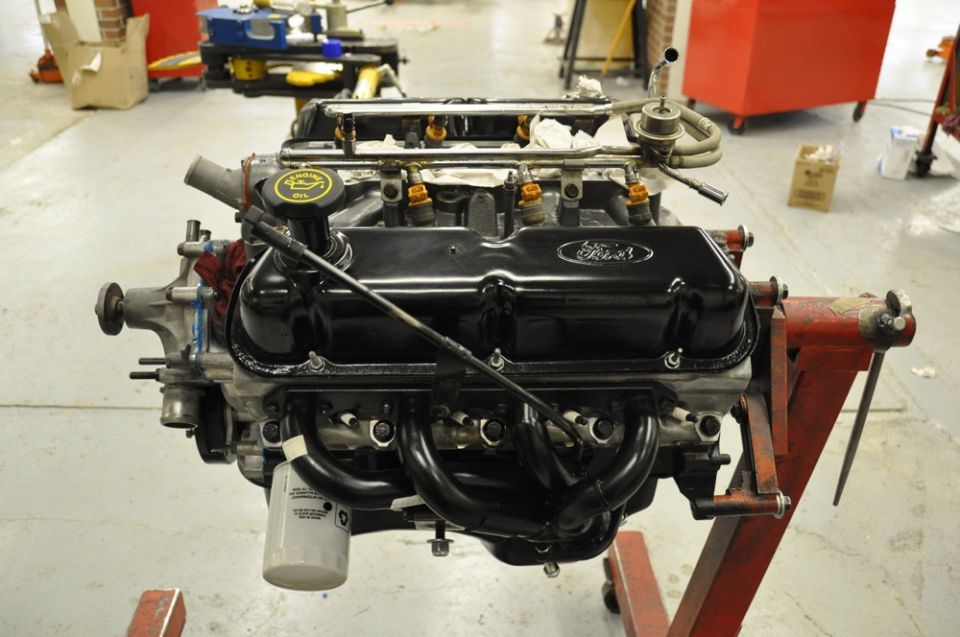 Ford V8 engine with black valve covers and exhaust headers, mounted on a red engine stand