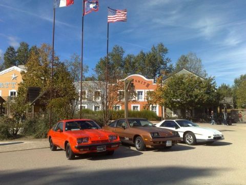 1991 Ford LTD Crown Victoria - Three vintage sports cars are parked in front of buildings with American flags flying overhead.