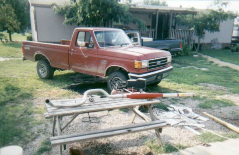 1987 Ford F-150 - A red 1987 Ford F-150 pickup truck is parked in a yard, with a picnic table holding exhaust parts in the foreground.