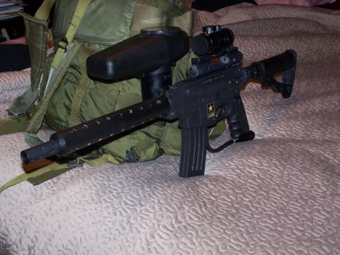 1986 Ford Bronco - A paintball marker with an attached hopper and scope rests on a bed next to a military-style backpack.