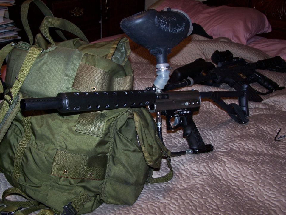 Green duffel bag with a paintball gun and hopper resting on a bed