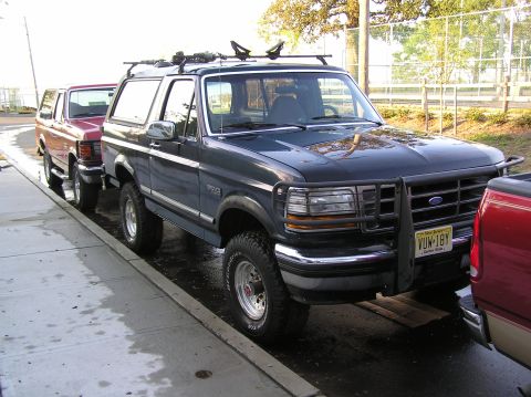 1993 Ford Bronco - A dark blue Ford Bronco is parked on the street, with a red vehicle partially visible in the foreground and another red vehicle behind it.