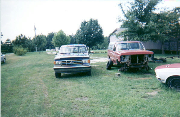 Dark Ford pickup parked beside a red and white pickup stripped to its bare front end in a yard