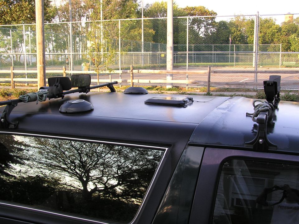 Roof of dark Ford Bronco with rack crossbars, rope, vents and roof panel, fence and trees behind