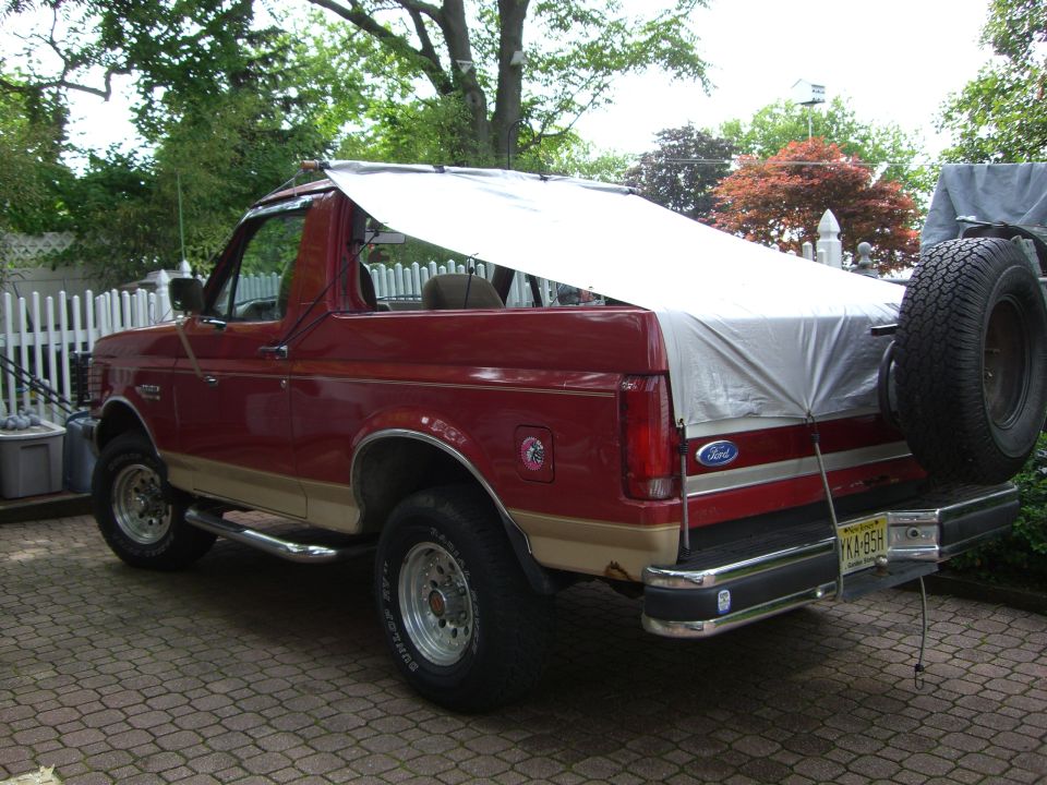 Red and tan Ford Bronco SUV with top off, gray tarp draped over rear, parked on brick driveway