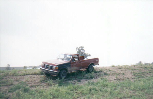 Faded red Ford F-Series long bed pickup parked on a grassy hill, distant driver side view