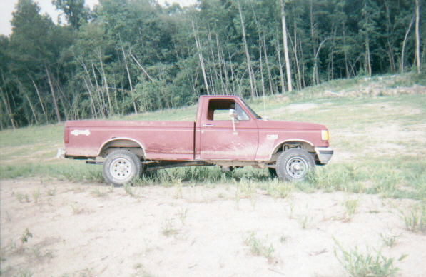 Mud-splattered dark red Ford long bed pickup parked on sand, passenger side view