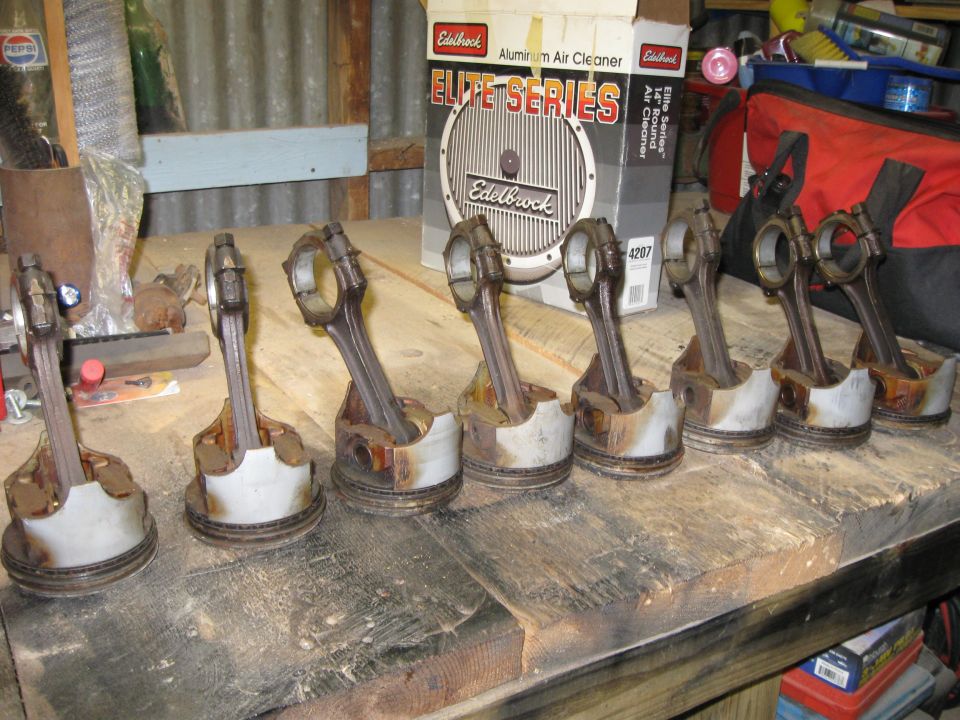 Eight used pistons with connecting rods arranged on a wooden workbench, with an Edelbrock air cleaner box in the background