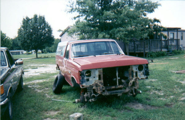 Red and white Ford pickup stripped of its grille and front end, sitting frame-down in a yard
