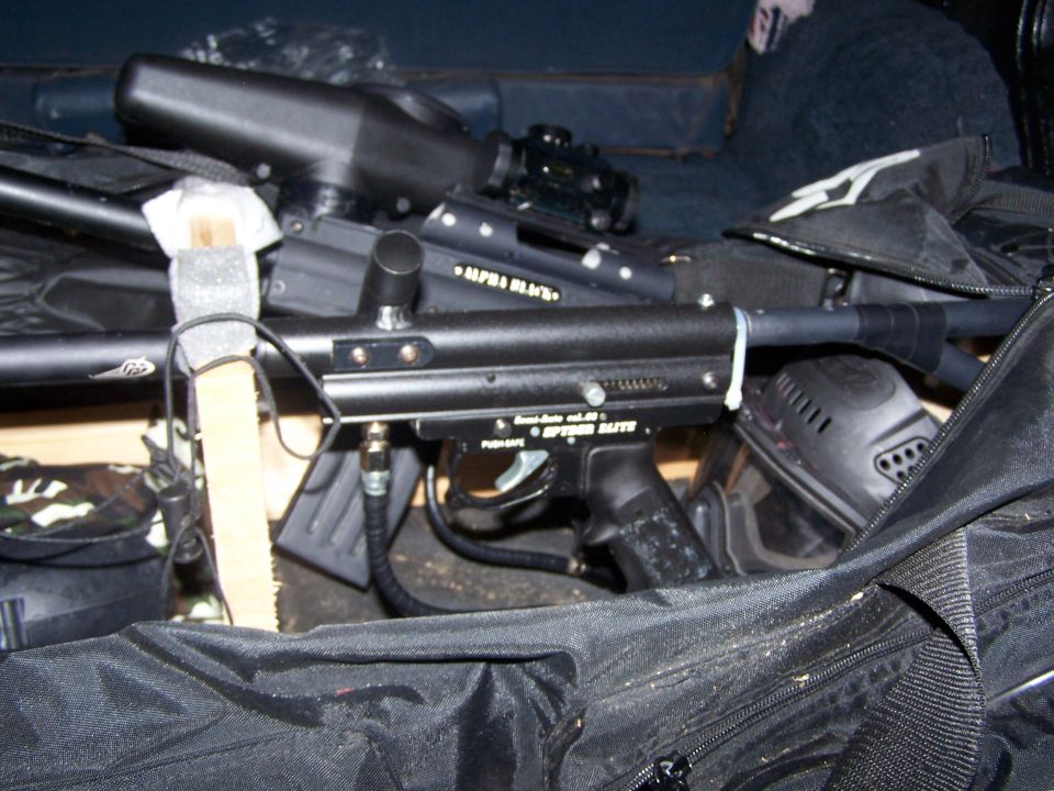 Paintball marker with "ALPHA BLACK" and "SPEEDER ELITE" visible, next to a mask and gear bag