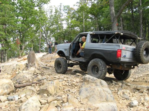 1990 Ford Bronco - Primer-gray topless Ford Bronco crawling over a rocky ridge trail, shown from the rear three-quarter with a full exterior roll cage and rol…