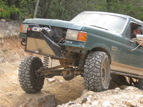 1990 Ford Bronco - Green Ford Bronco articulating over rocks on a trail, front axle drooped with the driver-side coil spring visibly unseated from its perch.…
