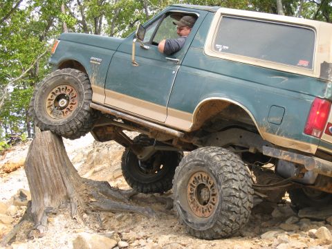 1990 Ford Bronco - Green and tan two-tone Ford Bronco flexing on a rocky trail, front driver tire climbing a weathered tree stump while the spare tire hangs o…