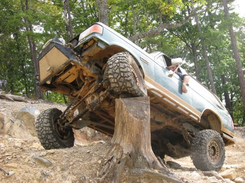 1990 Ford Bronco - A mud-caked green Ford truck flexes hard with its front end climbing a tree stump on a rocky wooded trail, front driver tire drooped and th…