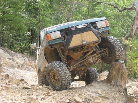 1990 Ford Bronco - A mud-covered green Ford 4x4 climbs a tree stump on a rocky trail, the passenger front tire lifted high while the driver side tucks, exposi…