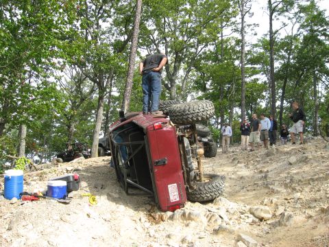 1990 Ford Bronco - A red 4x4 with an exposed roll cage and mud tires lies rolled onto its driver side on a rocky ridge trail, tailgate and license plate facin…