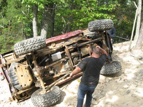 1990 Ford Bronco - A red 4x4 lies on its side on a rocky wooded trail, its underside fully exposed showing the frame rails, muffler, transfer case, leaf sprin…