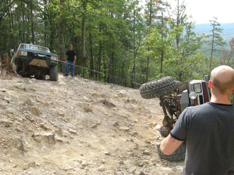 1990 Ford Bronco - A green SUV with a winch bumper anchors a taut winch line across a rocky wooded trail to a rolled-over rig lying on its side at right, mud…