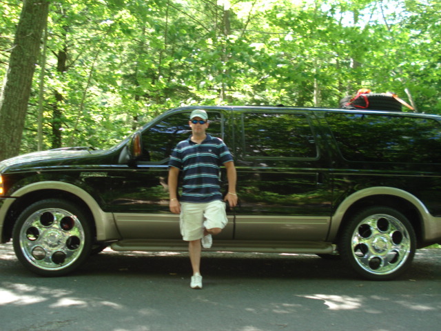 Man leaning against side of black and tan Ford Excursion with chrome wheels on wooded road
