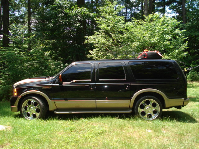 Black Ford Excursion with tan lower body panels, chrome wheels, and a spare tire on the roof rack, parked on grass with trees in the background