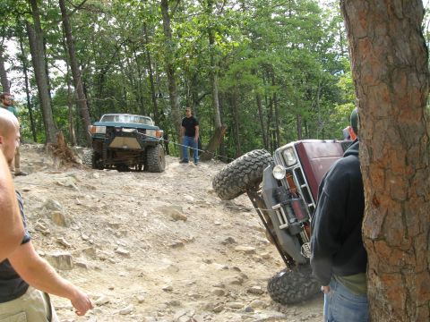 1990 Ford Bronco - A red truck lies tipped on its side against a tree on a rocky wooded trail, its winch and front bumper facing the camera, while a green SUV…